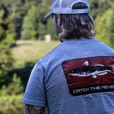 Rear view of a person wearing a grey Catfish short sleeve lifestyle T shirt displaying a large catfish graphic with the Catch The Fever® logo, standing near a pond in an outdoor setting.