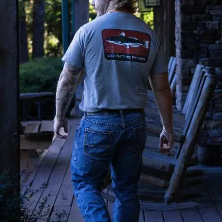 Rear view of a person walking on a wooden porch wearing a grey Catfish short sleeve lifestyle T‑shirt featuring a large catfish graphic and Catch The Fever® logo.