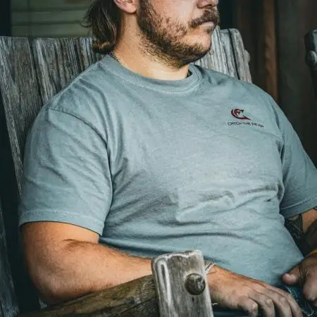 Catch The Fever® Marketing Director Dillon Weaver wearing a grey Catfish Short Sleeve Lifestyle T‑shirt with a small maroon logo, seated on a rustic chair.