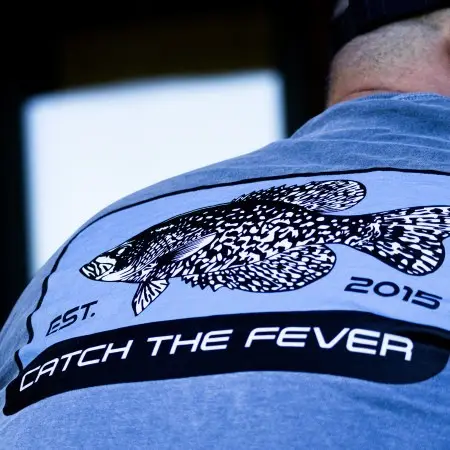Close-up of the back design on a dark gray Crappie short sleeve lifestyle T shirt, featuring a detailed black and white crappie fish graphic, 'EST. 2015' text, and the Catch The Fever® logo.