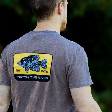 Rear view of a man wearing a grey Crappie short sleeve lifestyle T shirt with a yellow crappie fish graphic and Catch The Fever® logo on the back, standing outdoors.