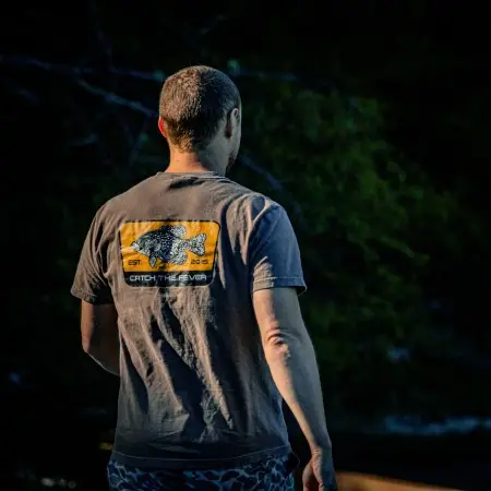 Rear view of a man wearing a brown Crappie short sleeve lifestyle T shirt with a yellow crappie fish graphic and Catch The Fever® logo, standing near water at dusk.