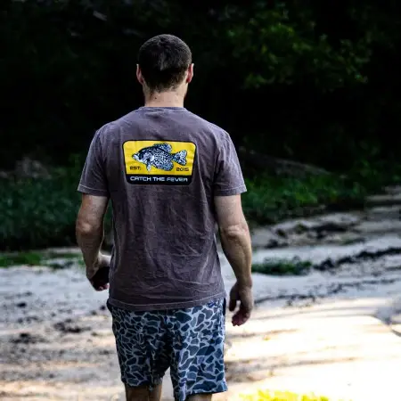Rear view of a man walking along a sandy path wearing a brown Crappie short sleeve lifestyle T shirt featuring a large yellow crappie fish graphic and Catch The Fever® logo.