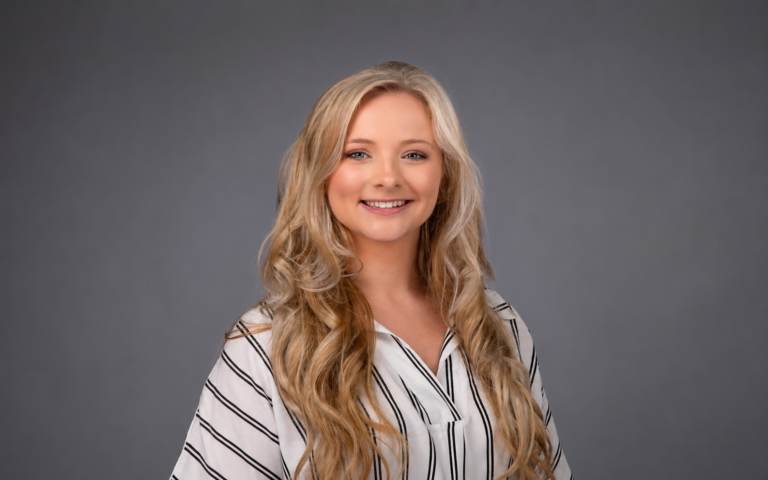 Smiling young woman with long blonde wavy hair wearing a white striped blouse against a gray studio backdrop