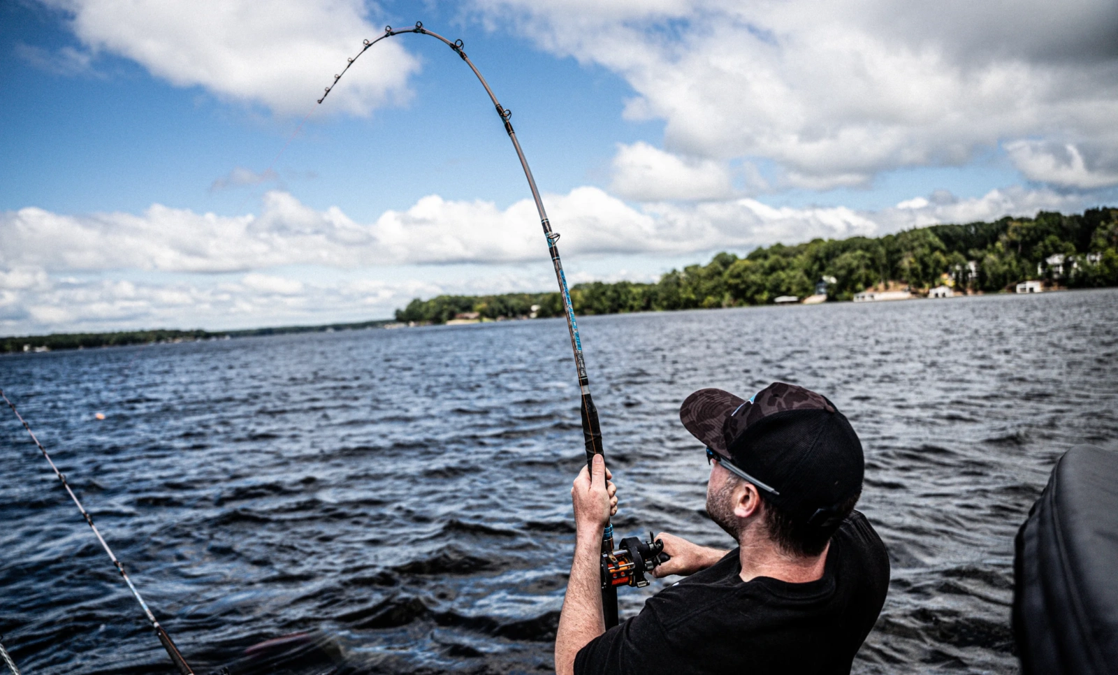 Angler leaning back on a deeply bent Catch The Fever rod while fighting a fish on open lake water under a bright summer sky.